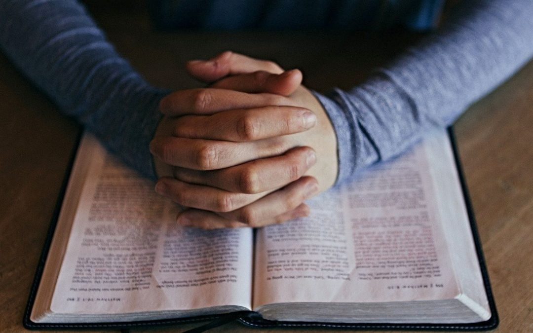 Crossed hands resting on pages of open Bible