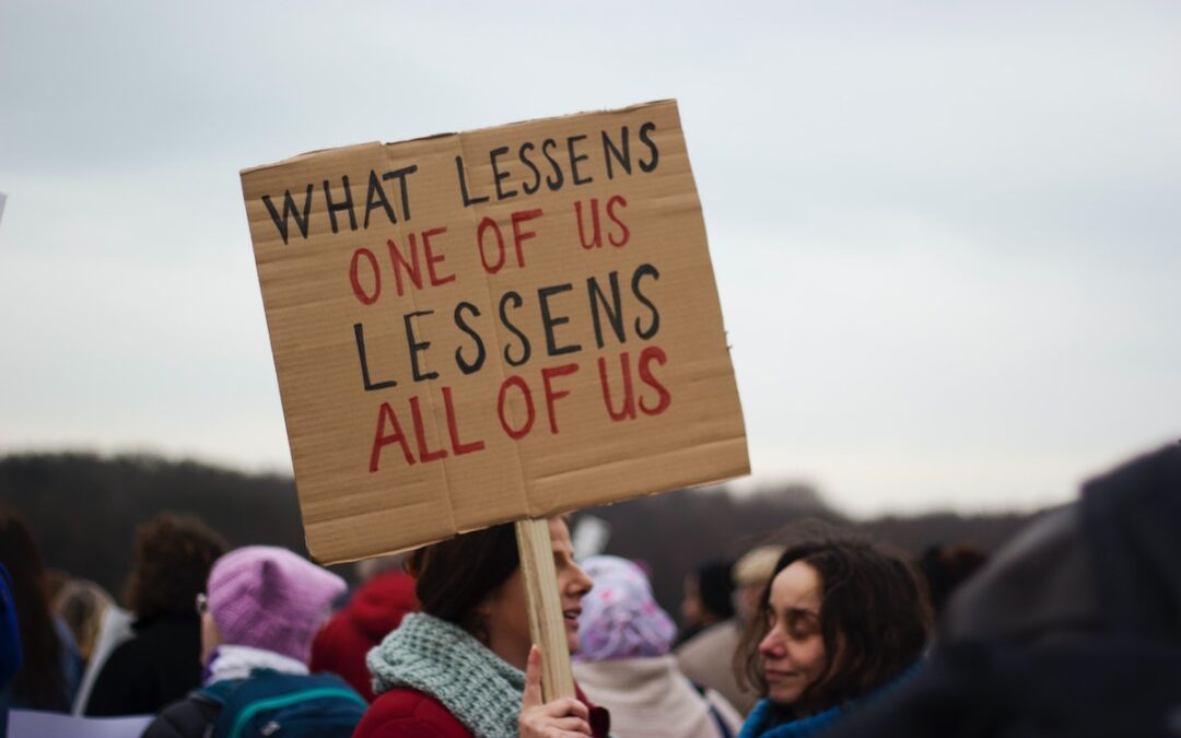 A woman holding a cardboard sign that says, “What lessens one of us lessens all of us.”