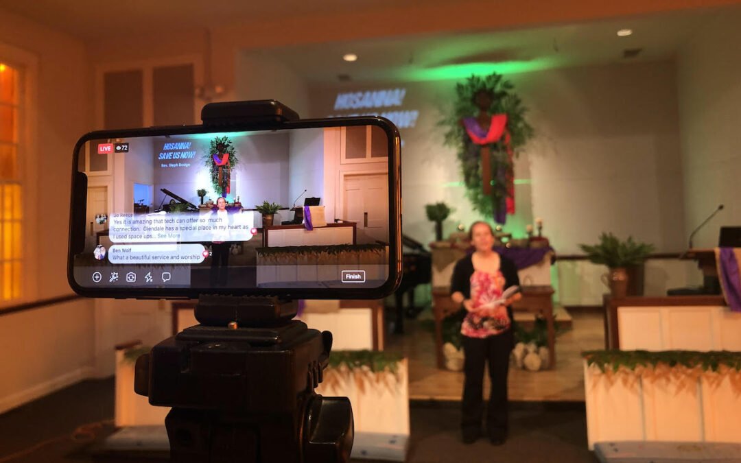 A smartphone in the foreground livestreaming a pastor standing alone in a church deliver a sermon.