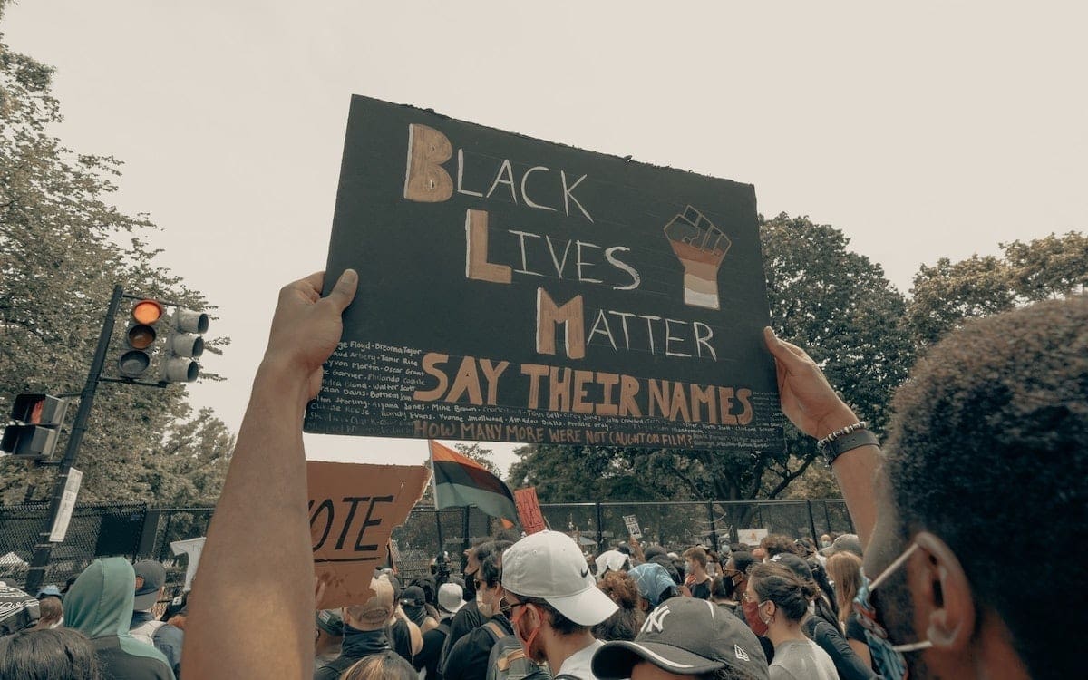 A man at a protest holding up a sign that says, among other things, “Black Lives Matter,” “Say Their Names” and “How Many More Were Not Caught on Film?”