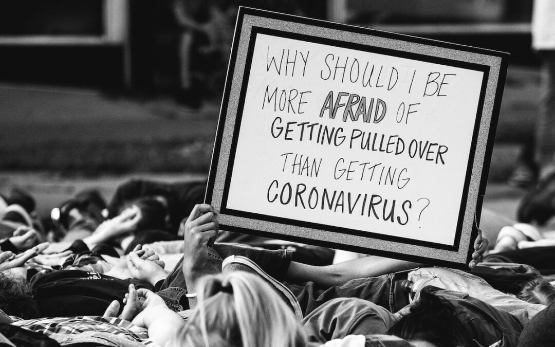 Protestors laying on the ground with one holding up a sign that says, “Why should I be more afraid of being pulled over than getting the coronavirus?”