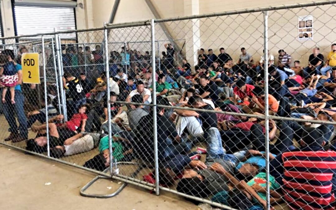 Immigrants crowded in a U.S. immigration facility behind a chain-link fence.