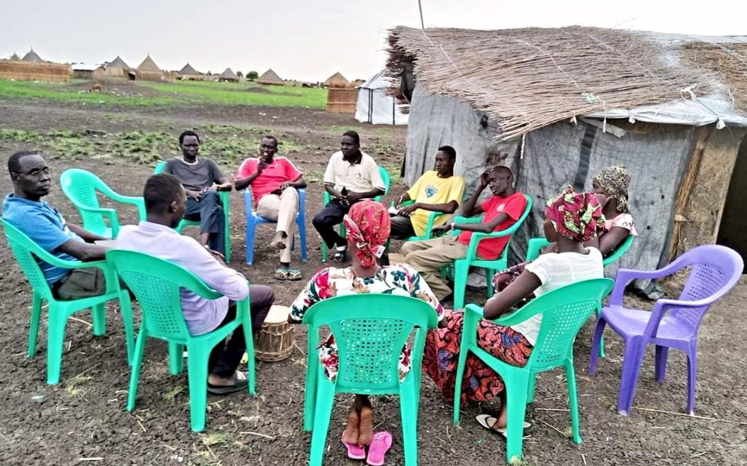 A gathering of refugees who returned to South Sudan sitting in green chairs in a circle outside.