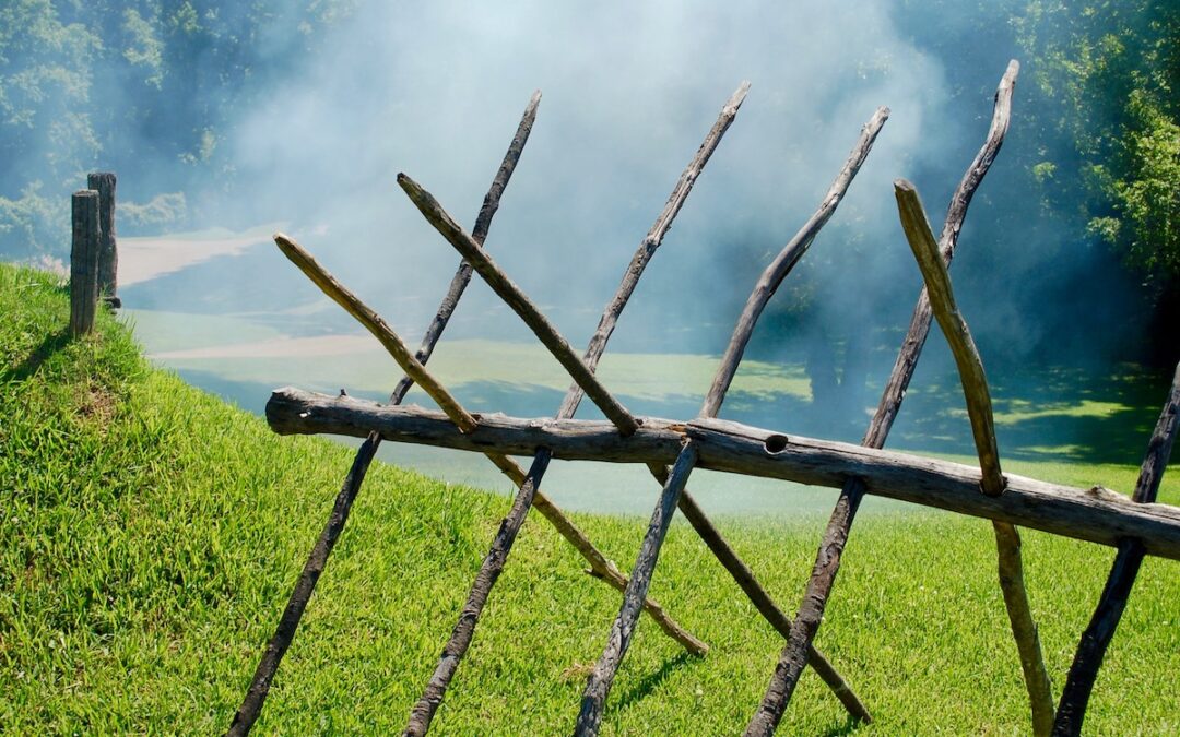 A grassy field obscured with smoke and a stick barricade in the foreground.