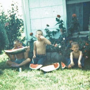 Three boys sitting outside barefoot eating watermelon.