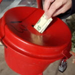 A red Salvation Army donation kettle with someone placing a $10 bill inside.
