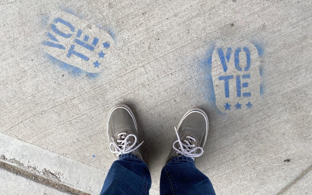 A person’s feet seen from above on a sidewalk next to the word “Vote” spray painted in blue.