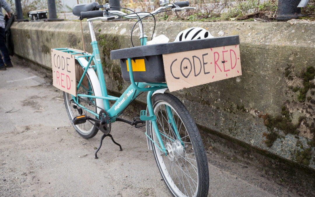 A bike parked on a sidewalk with the words “code red” on cardboard signs attached to the front and rear of the bike.