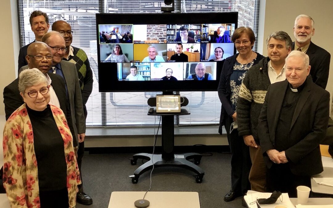 A group of people standing on both sides of a TV with several people in a live call on the screen.