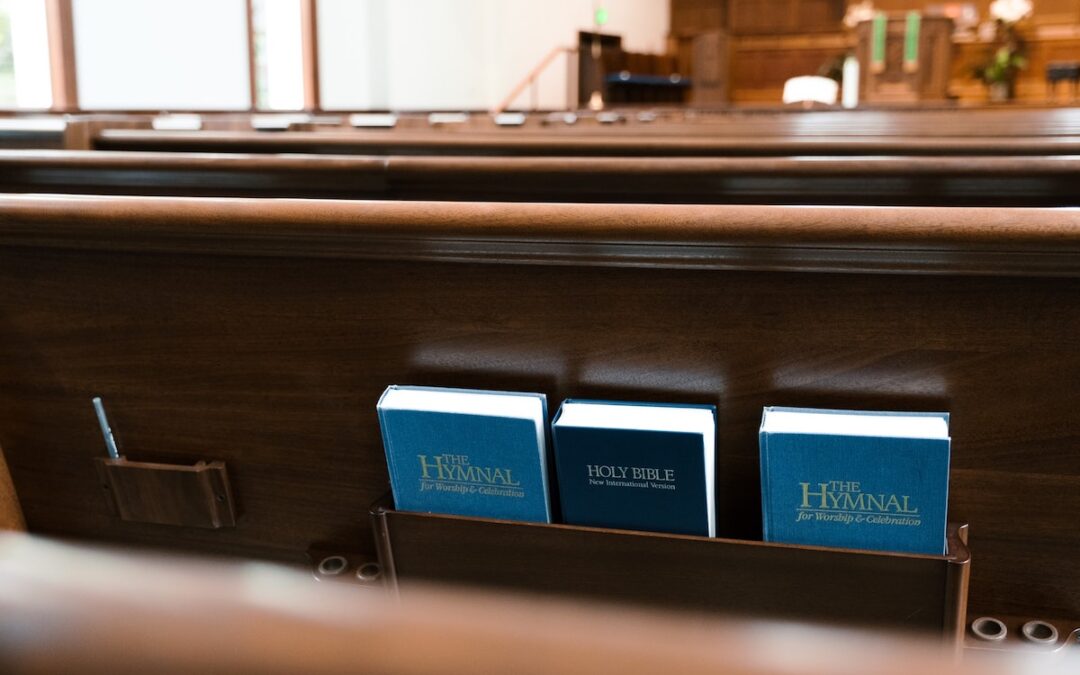Empty pews in a church worship space, with a Bible and two hymnals in a wooden holder on the back of one pew.
