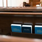 Empty pews in a church worship space, with a Bible and two hymnals in a wooden holder on the back of one pew.