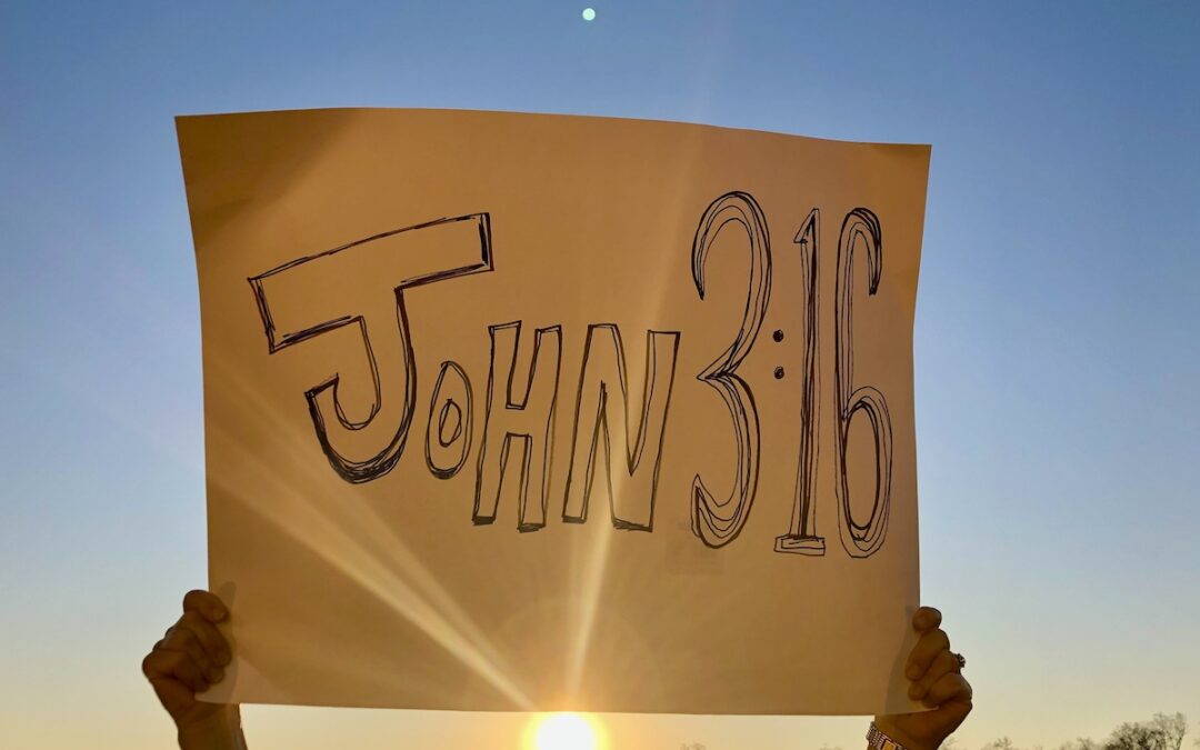 A yellow sign held in the air with John 3:16 written on it in black marker.