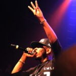 Kendrick Lamar holding a microphone in one hand and holding up three fingers on his other hand.