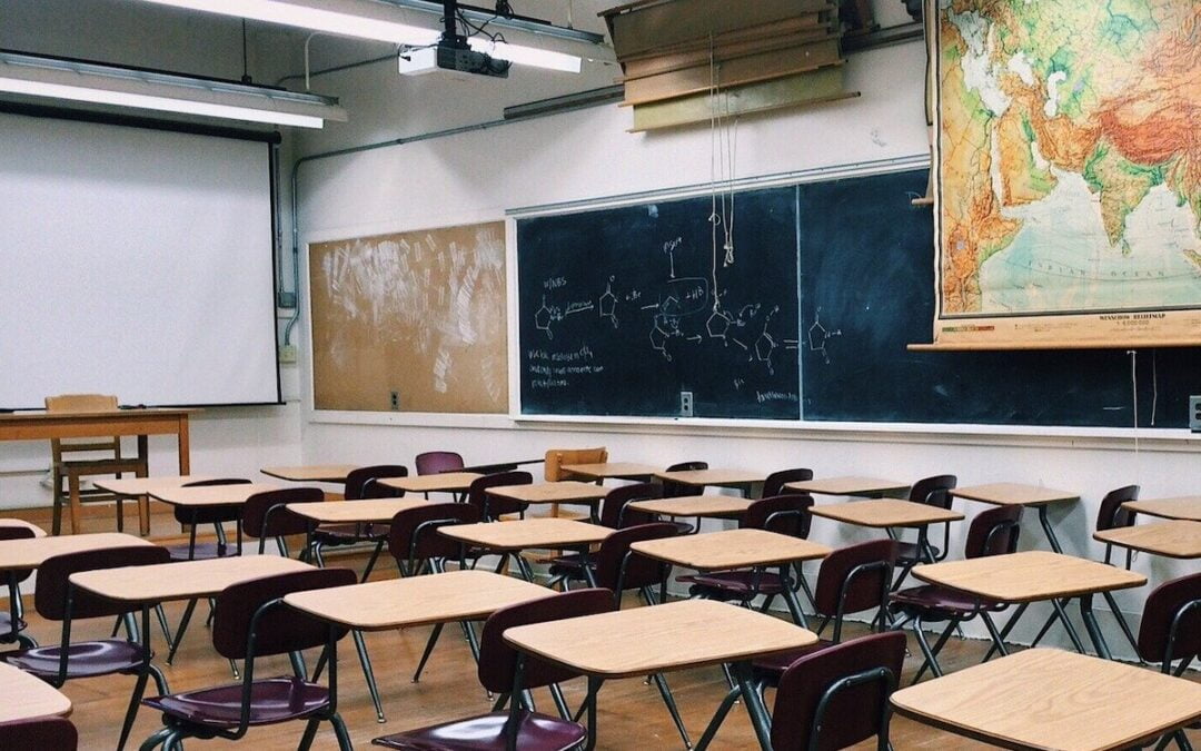 An empty school classroom with student desks and a teacher’s desk, along with chalkboards and maps on the wall.