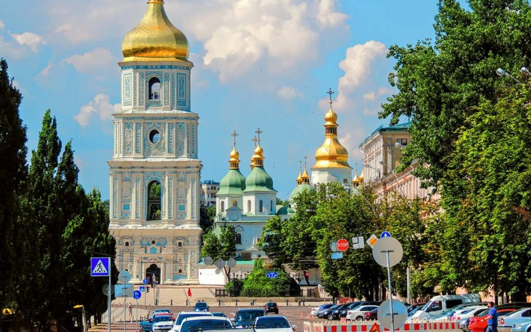 A view of the golden domes on the top of St. Sophia Cathedral in Kyiv, Ukraine, seen from a street, with a bright blue sky and white clouds in the background.