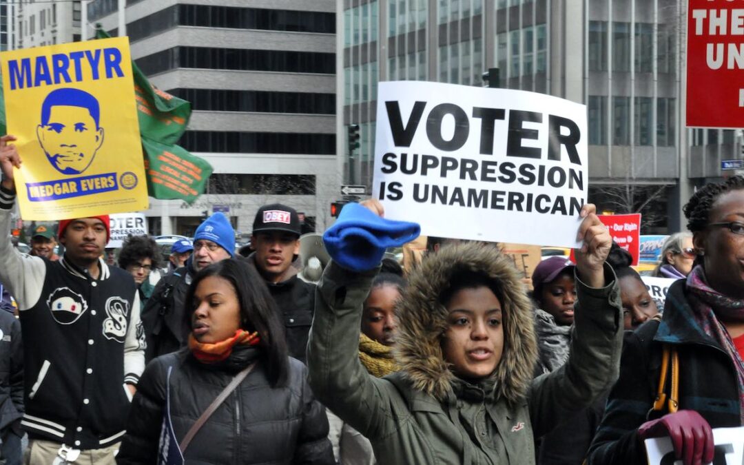 A group of people marching together on a street with one holding a white sign with black letters that says, “Voter suppression is unamerican.”