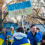 A man draped in a Ukrainian flag holds a “Stop the War” sign in a crowd.