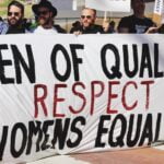 Several men standing side-by-side on the street holding a large, white sign that says, “Men of quality respect women’s equality.”