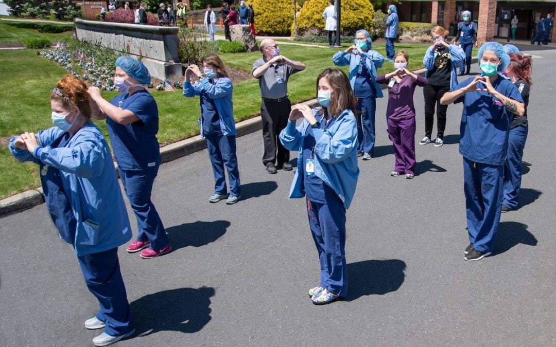 A group of nurses standing outside on a sidewalk making a heart shape with their hands.