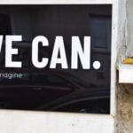 A black sign with white letter that says, “We can. Just imagine.”