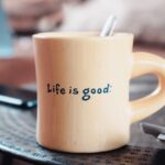 A coffee mug with the words “Life is Good” on the side sitting on a table.