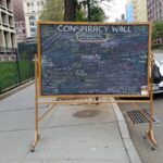 A chalkboard on the street filled with writing and with a title, “Conspiracy Wall.”