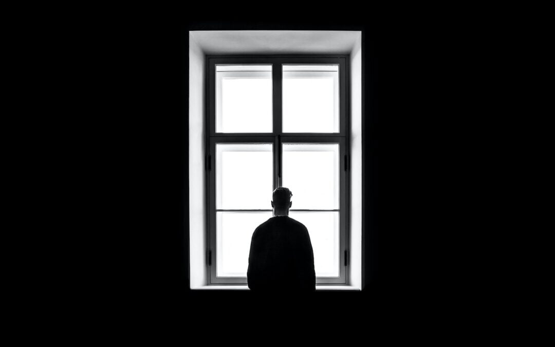 A man seen from behind standing in a black room looking out a window.