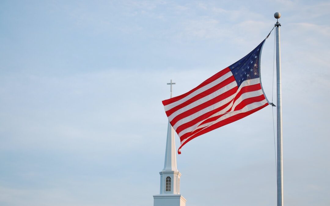 A U.S. flag with a church steeple with a cross in the background set against a blue sky.