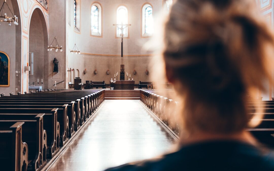 The back of a woman’s head out of focus in the foreground looking into an empty church sanctuary seen in focus in the background.
