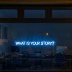 A neon sign on a storefront window that asks, “What is your story?”