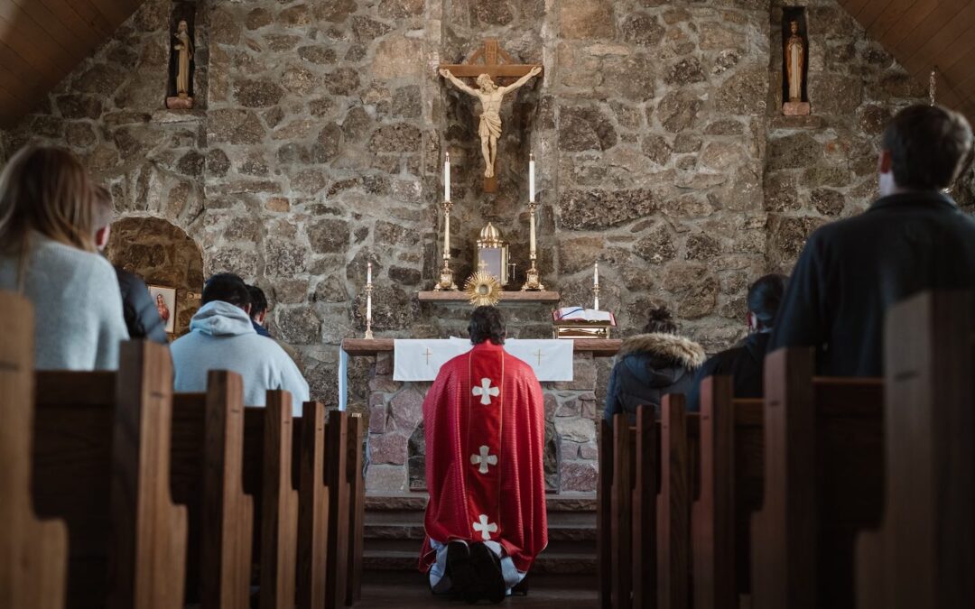 The interior of a Catholic church during Mass seen from the back with parishioners sitting and looking at a priest kneeling at the altar.