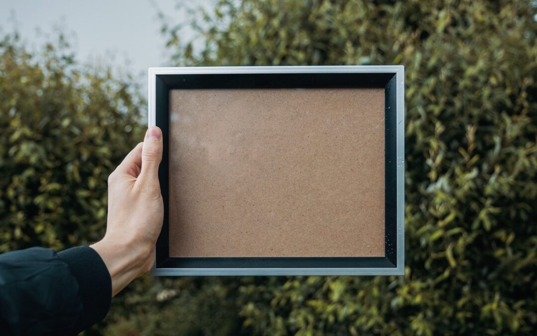 A person’s hand holding an empty picture frame outside with trees in the background.