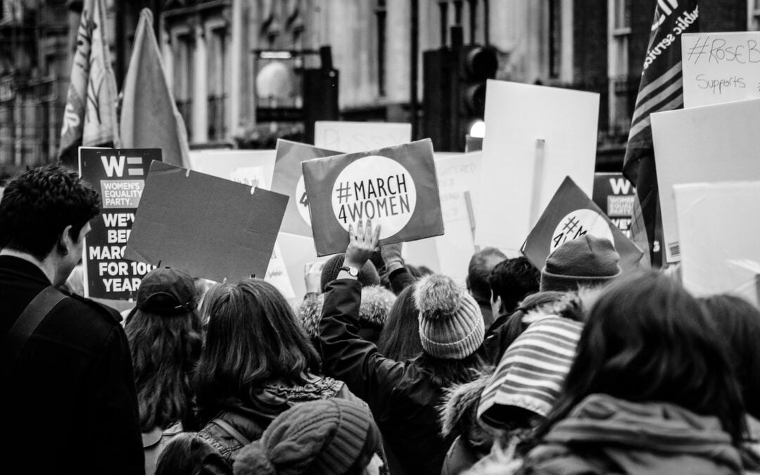 A black-and-white image of a group of people seen from behind holding signs in a demonstration for women’s equality.