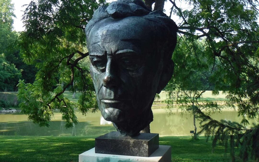 A metal bust of theologian Paul Tillich on a concrete base with a river or pond in the background.