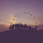 A silhouette view of a group of graduates wearing their gowns and throwing their hats in the air at sunset.