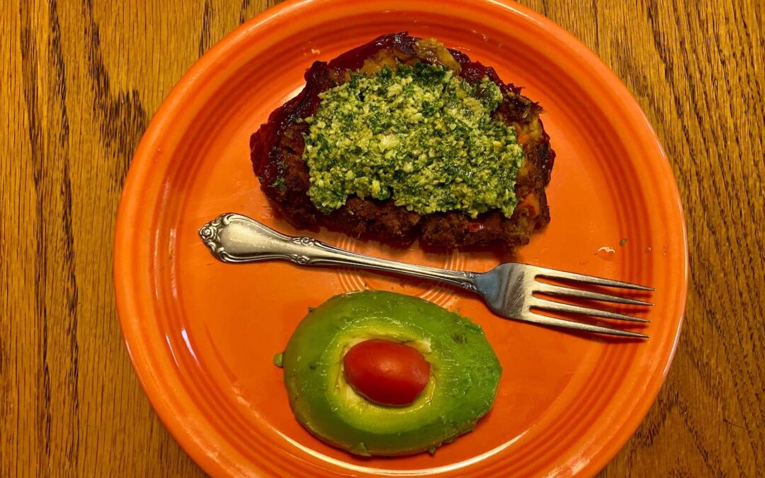 An orange plate with a silver fork in the middle and a piece of bread with basil pesto on it and below the fork an avocado with a small tomato in the middle.