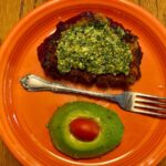 An orange plate with a silver fork in the middle and a piece of bread with basil pesto on it and below the fork an avocado with a small tomato in the middle.