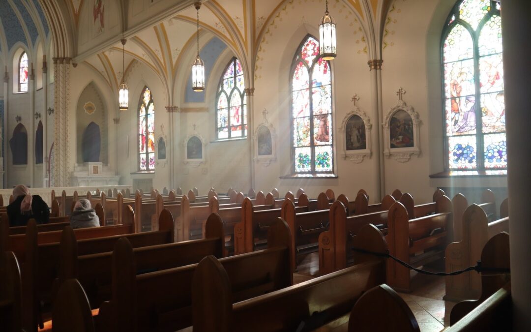 The interior of a church showing rows of mostly empty pews with the sun shining through several stained-glass windows.