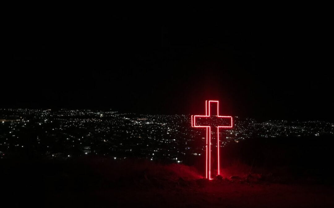A neon cross lit up at night on a hillside overlooking a town with lights from houses and business visible in the distance.