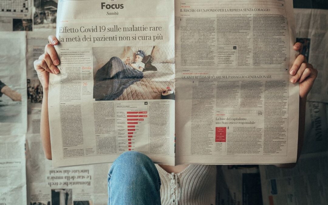 A person in blue jeans sitting on the floor holding up a newspaper, with other newspapers on the wall behind them.