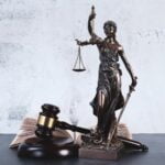 A metal statue of “lady justice” on a table next to a gavel and an open book.