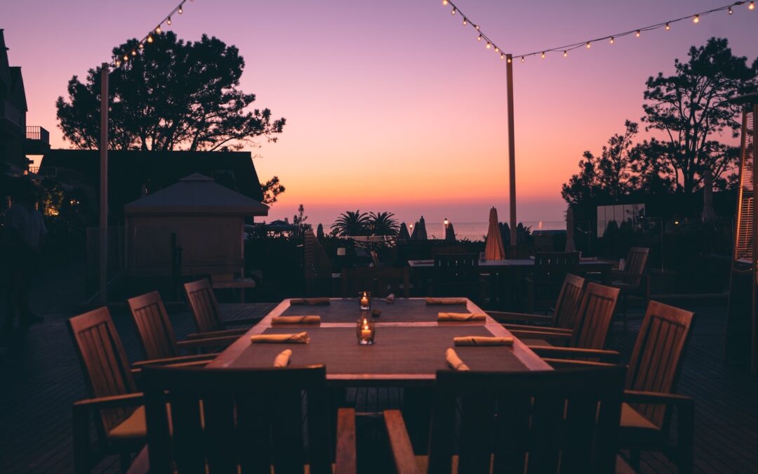 A table on a patio outside with chairs around it and napkins on the top, seen in the foreground with the sun setting in the background.