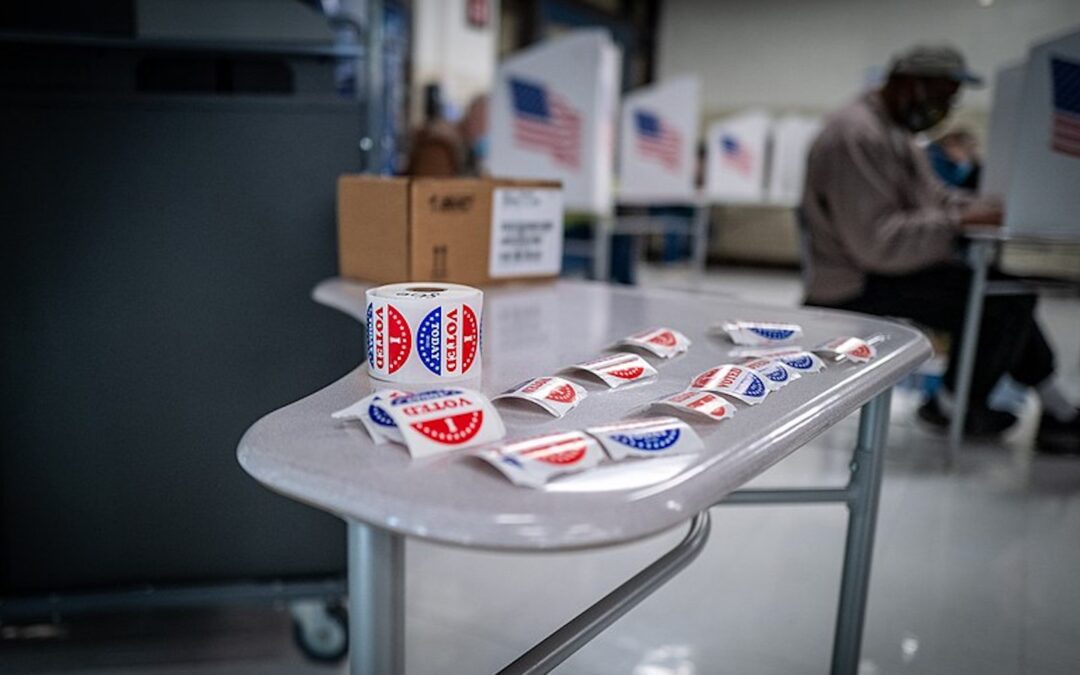 A table with “I voted” stickers on a table in the foreground with people sitting at voting booths casting their ballots in the background.