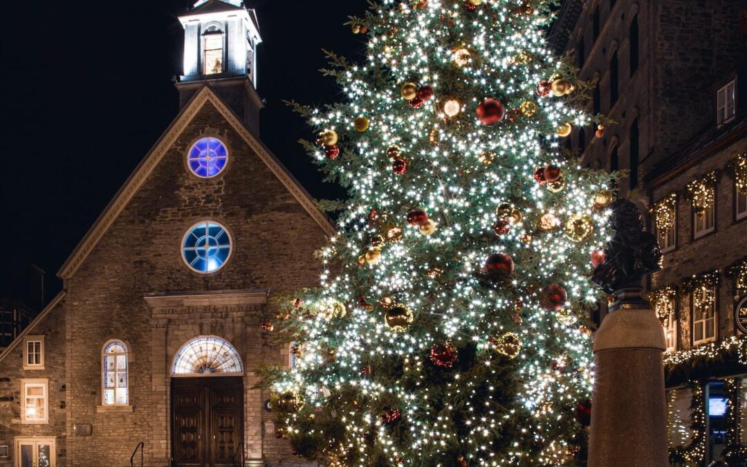 A large Christmas tree with lights and ornaments outside on a street with a church in the background.