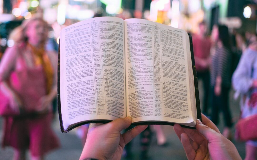 A person’s hands seen up close holding an open Bible on a busy sidewalk with passersby in the background out of focus.