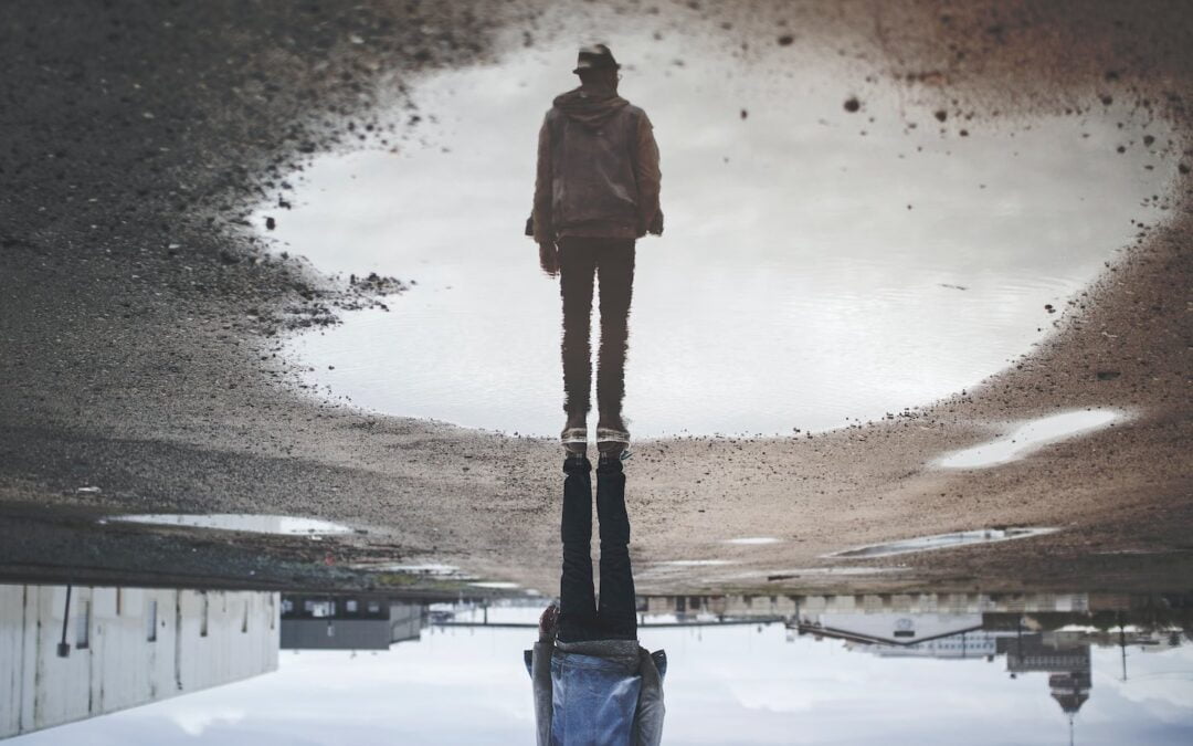 A man’s reflection in a pool of water on an asphalt road.