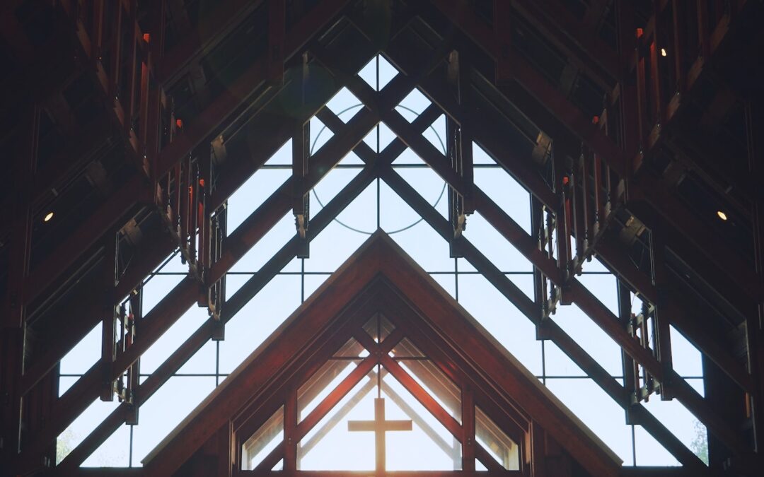 A church interior, showing the open sky through a large window and a wooden cross among the ceiling rafters.