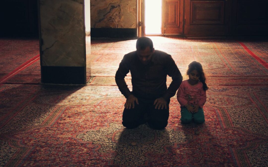 A father and daughter kneeling side by side on a rub praying in a mosque.
