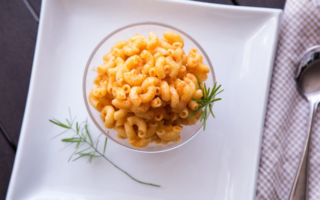 A clear bowl filled with macaroni and cheese seen from above sitting on top of a white plate on a table.