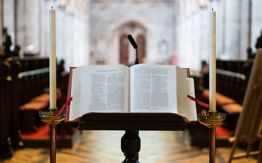 A Bible open on a wooden pulpit stand with candles on either side and an empty sanctuary out of focus in the background.